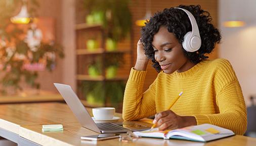 Smiling black girl with wireless headset studying online, using laptop at cafe, taking note